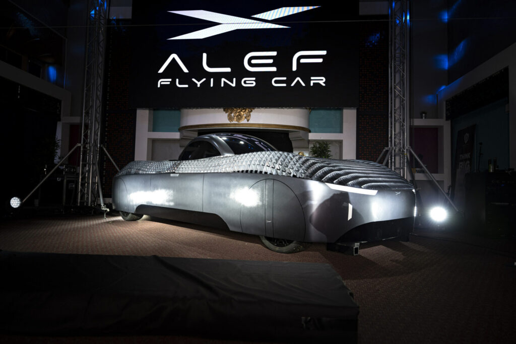 The alef flying car is on display in a room, featured on synthetic television news.