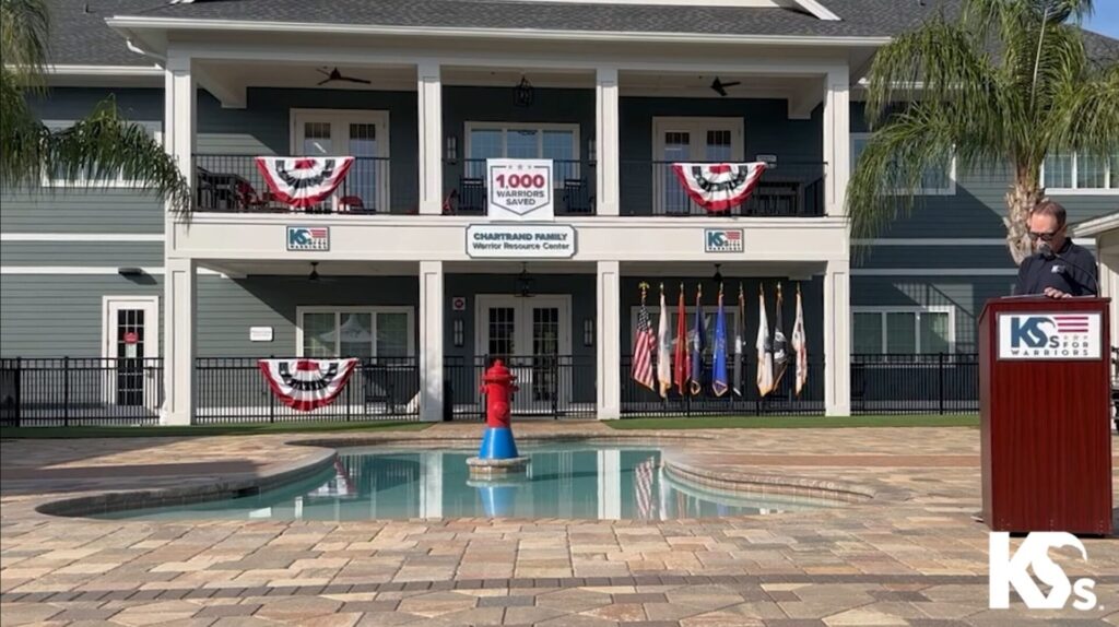 A person stands at a podium beside a swimming pool in front of a building adorned with patriotic decorations and multiple flags, celebrating a milestone achievement for K9s For Warriors.