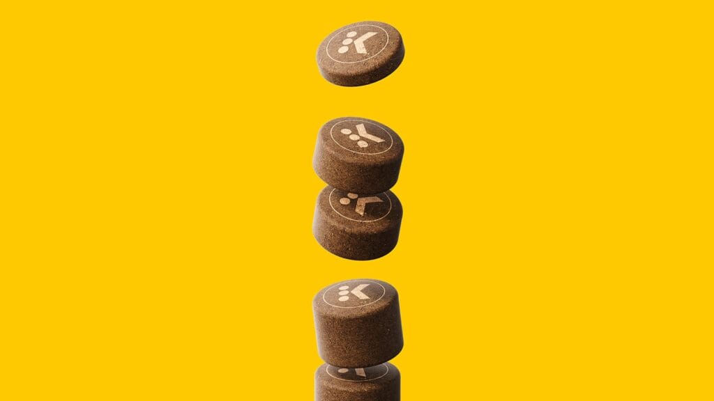 Five brown, cylindrical Keurig capsules with white symbols depicted on each, appear suspended in mid-air against a yellow background, arranged in a vertical line with one capsule separated from a stack of four.