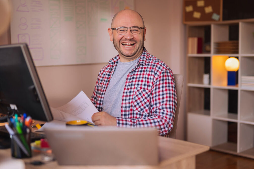 A cheerful bald man wearing glasses and a plaid shirt smiling at the camera while sitting at a desk with a laptop open to Accounting Software from Wave Apps and holding papers.
