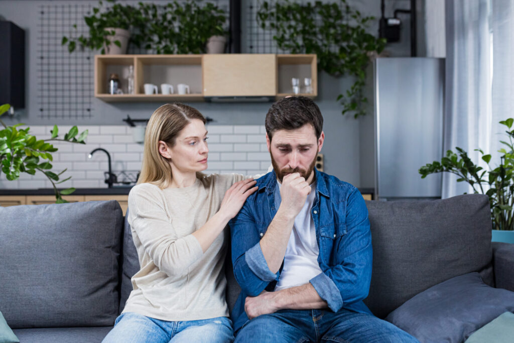 A woman comforting a distressed man as they sit together on a couch in a living room, pondering the elusive American Dream.