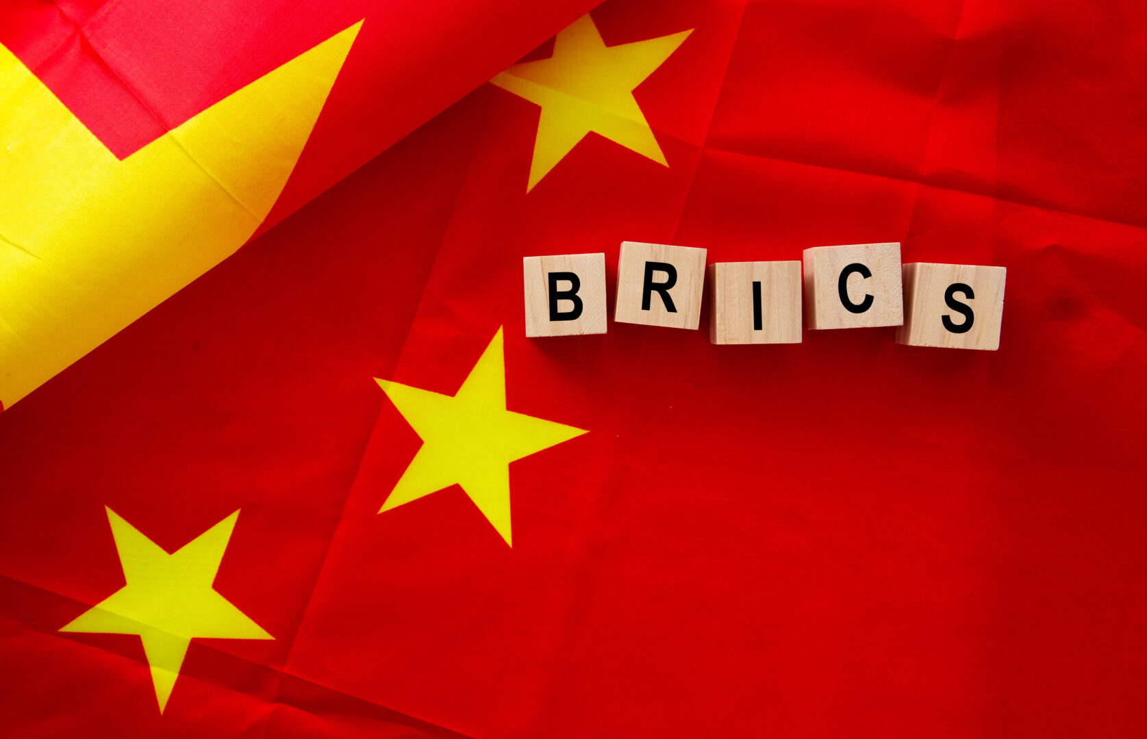 Wooden letter blocks spelling "Brics Alliance" on a red surface with a partial view of the Chinese flag visible.