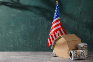 An American flag in a stand beside a cardboard house model and a rolled hundred dollar bill against a green textured background depict "The American Dream on Hold: Why Homes Cost More Than Your Dreams".