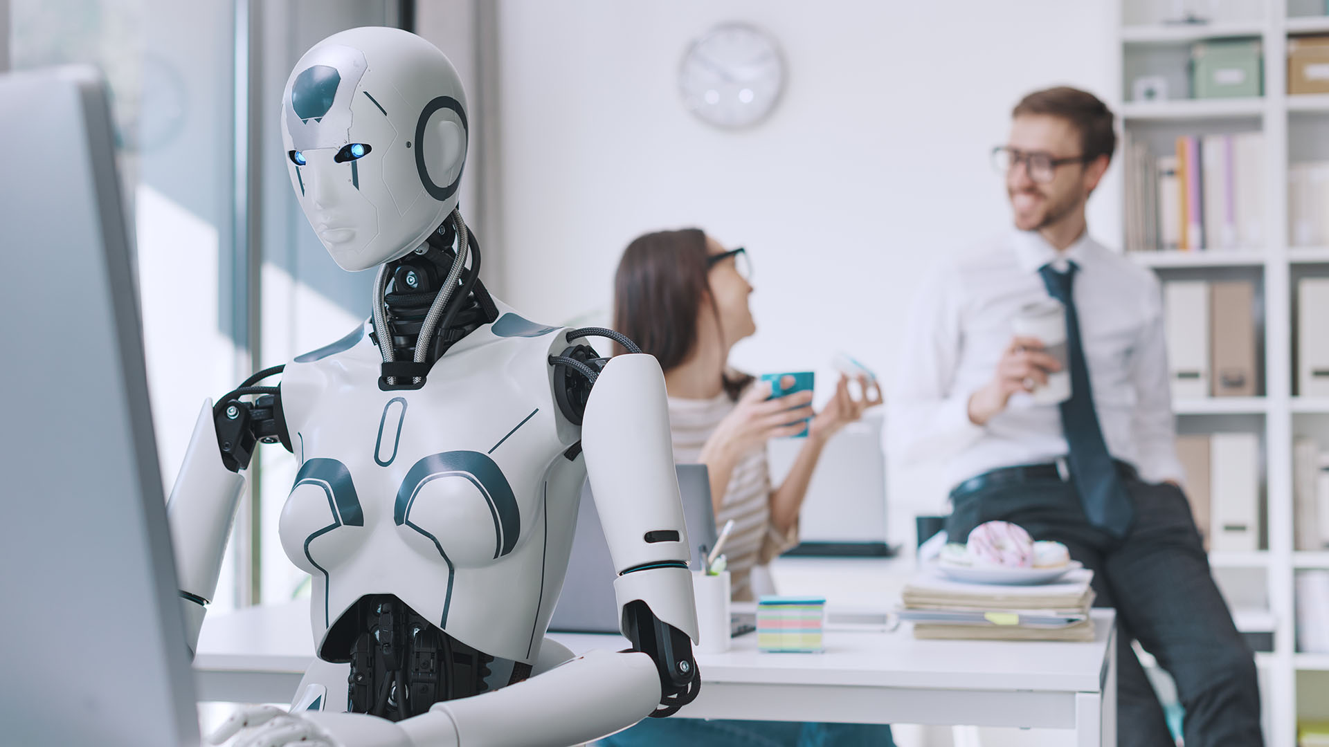 A humanoid robot works at a desk with a computer, while two people in the background converse and laugh in a modern office setting.