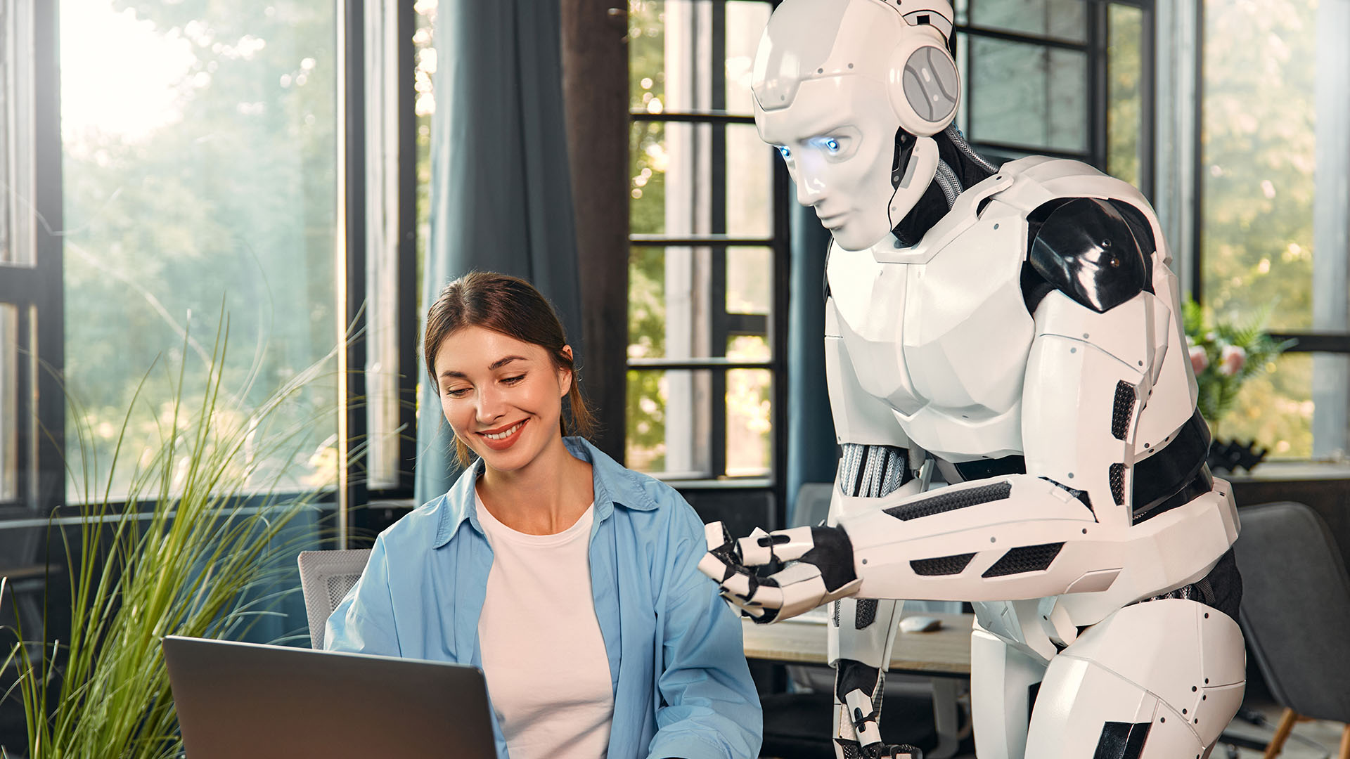 A woman, smiling, works on a laptop at a desk while a humanoid robot stands next to her, pointing toward the screen. They are in a modern room with large windows and green plants, setting the stage for the human vs. machine collaboration in this ever-evolving newsroom battle.