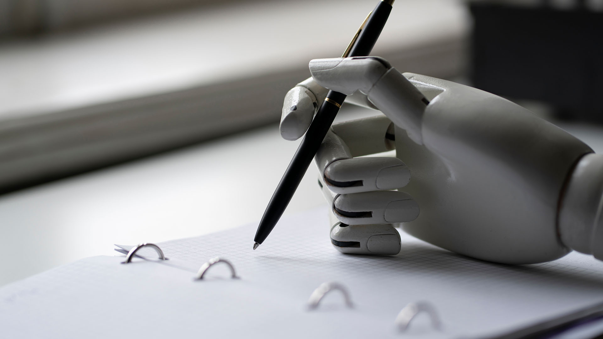 A robotic hand holds a pen over an open notebook with binder rings, symbolizing the ongoing Battle for the Newsroom as automation reshapes how stories are crafted.