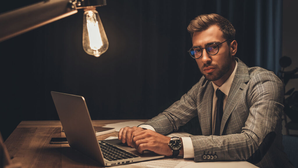 A man in a plaid suit and glasses sits at a wooden desk with a laptop, papers, and books under a hanging light bulb, looking at the camera, as if ready to engage in the ultimate battle between human vs. machine.