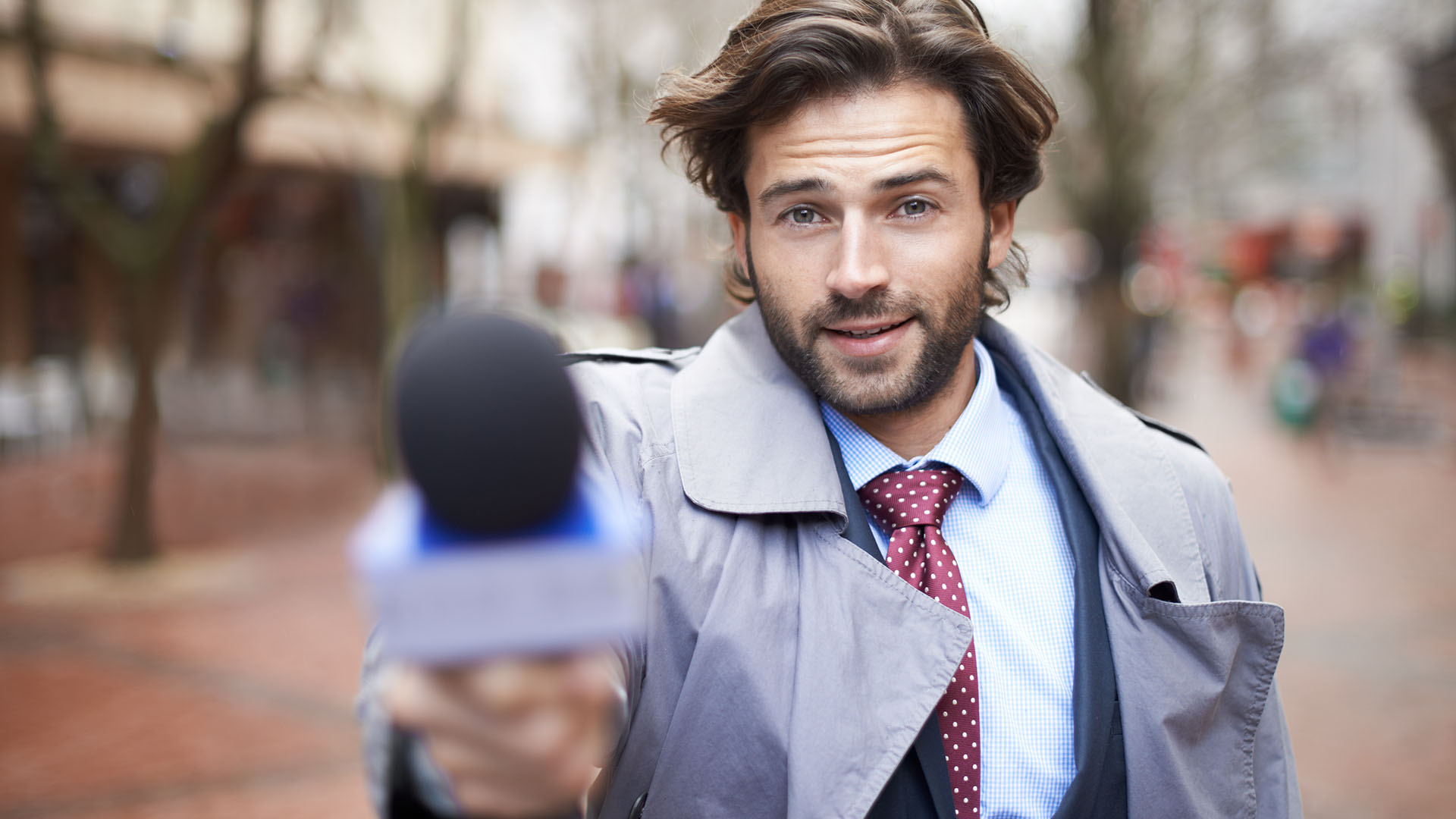 A man with long brown hair and a beard holds a microphone towards the camera. He is wearing a gray trench coat, blue shirt, and red tie, standing outdoors on a brick pavement street, ready for the Battle for the Newsroom.
