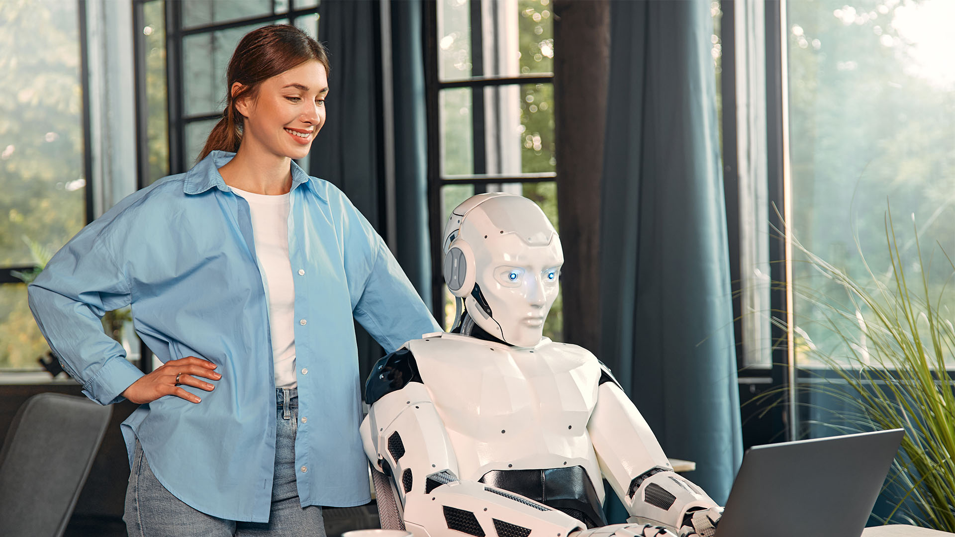 A woman in a blue shirt stands beside a seated humanoid robot looking at a laptop in a modern room with large windows, exemplifying the battle for the newsroom between human vs. machine.