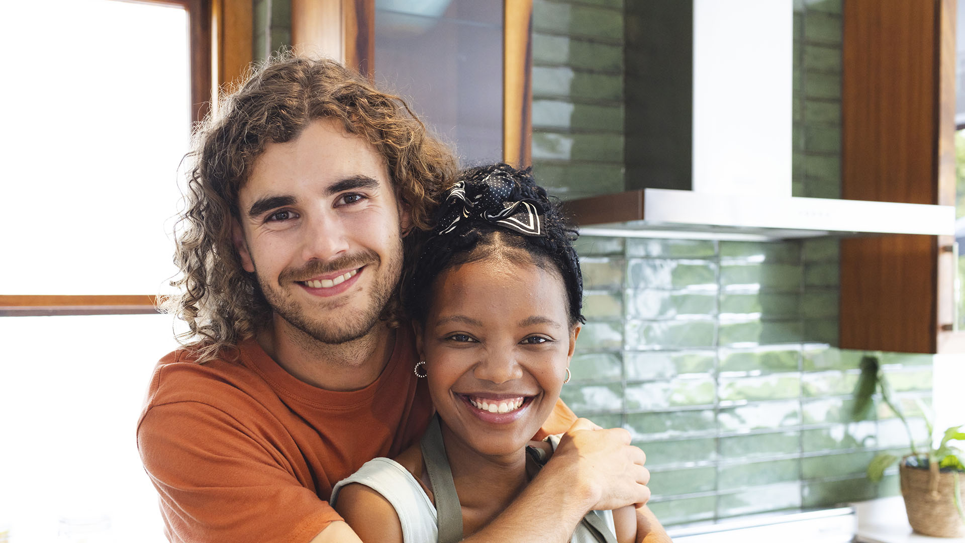 A man with curly hair and a woman with braids smile while hugging in a modern kitchen with green tiled walls, pondering if renting is ruining your future.