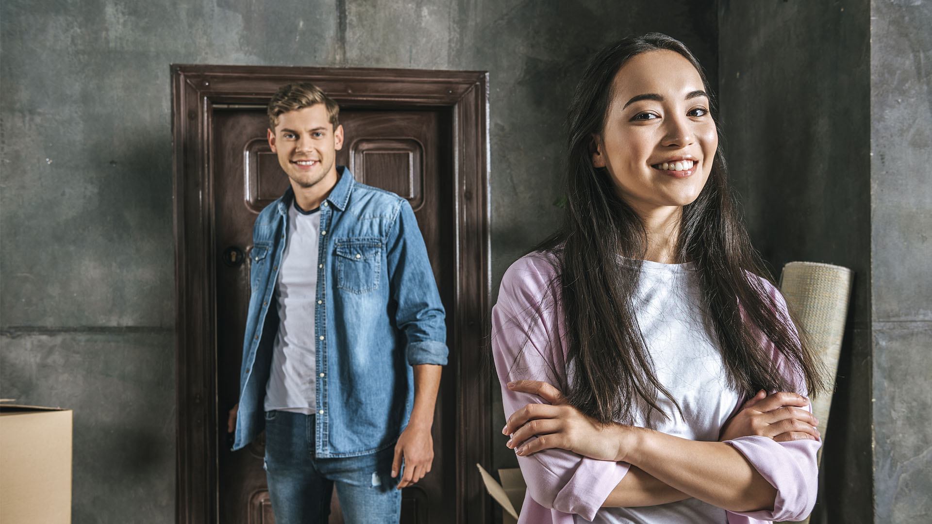 A woman with crossed arms smiles in the foreground while a man stands in the background near a door, both casually dressed in a room with gray walls. Is renting ruining your future? They seem to have found a comfortable balance.