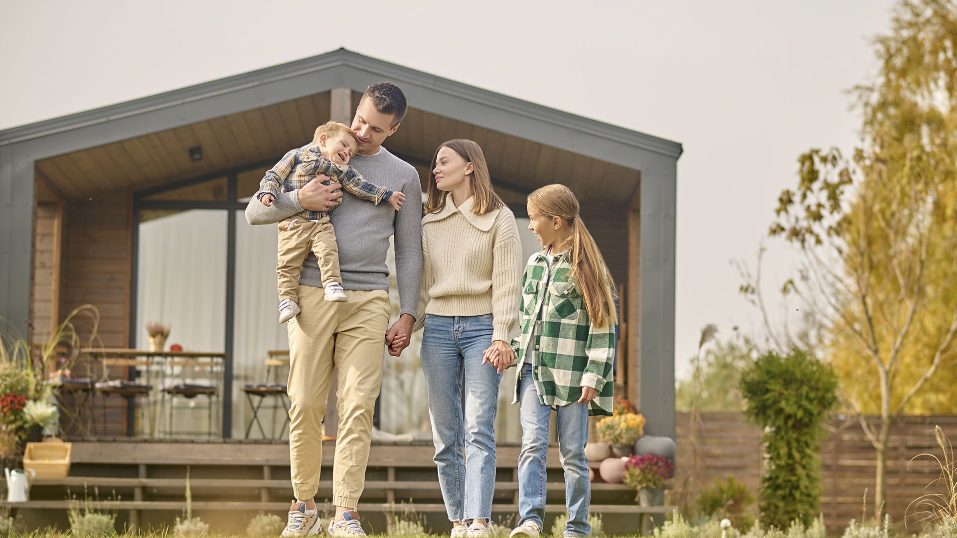 A family of four stands in front of a modern house, exemplifying the dream of homeownership. The father holds a toddler, the mother stands next to him holding his hand, and an older child stands beside them. Is renting ruining your future? Consider taking the next step towards owning your own home.