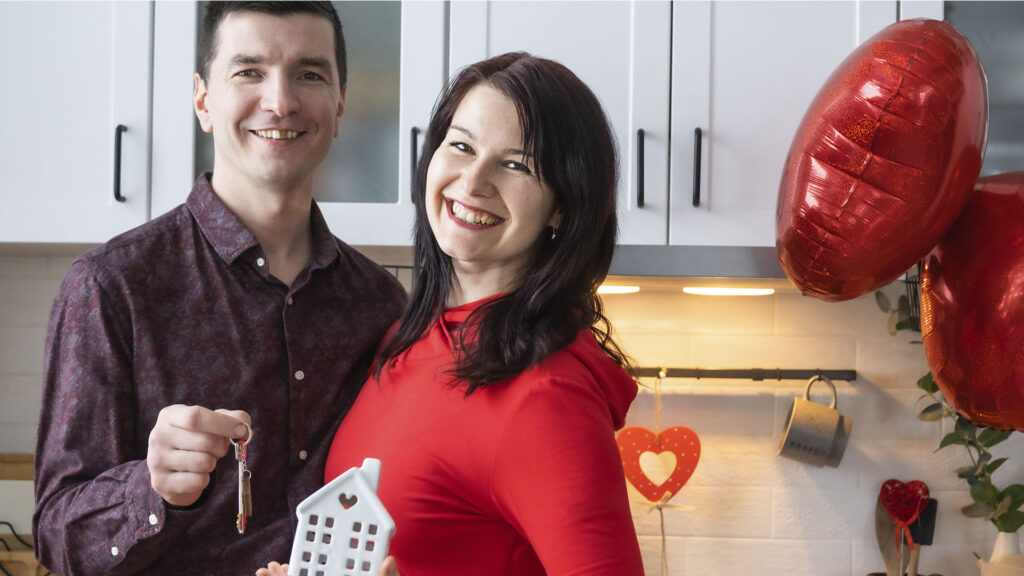 A man and woman smiling in a kitchen, holding a house key and a small house model. Red heart decorations hang in the background, perhaps hinting at the joy of homeownership. Is renting ruining your future?