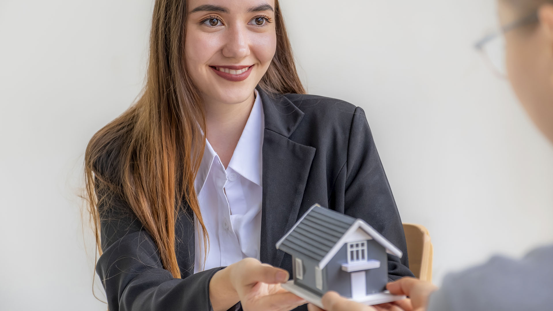 A woman in a blazer hands over a small model house to another person in front of a plain background, posing the question: Is renting ruining your future?