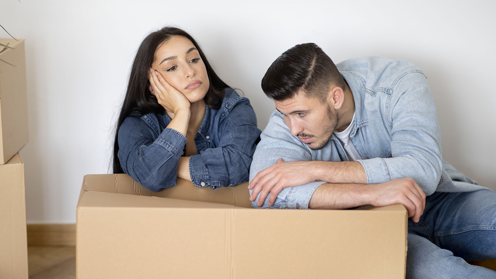 A woman and a man sit on the floor looking tired and contemplative, surrounded by cardboard boxes, pondering if renting is ruining their future.