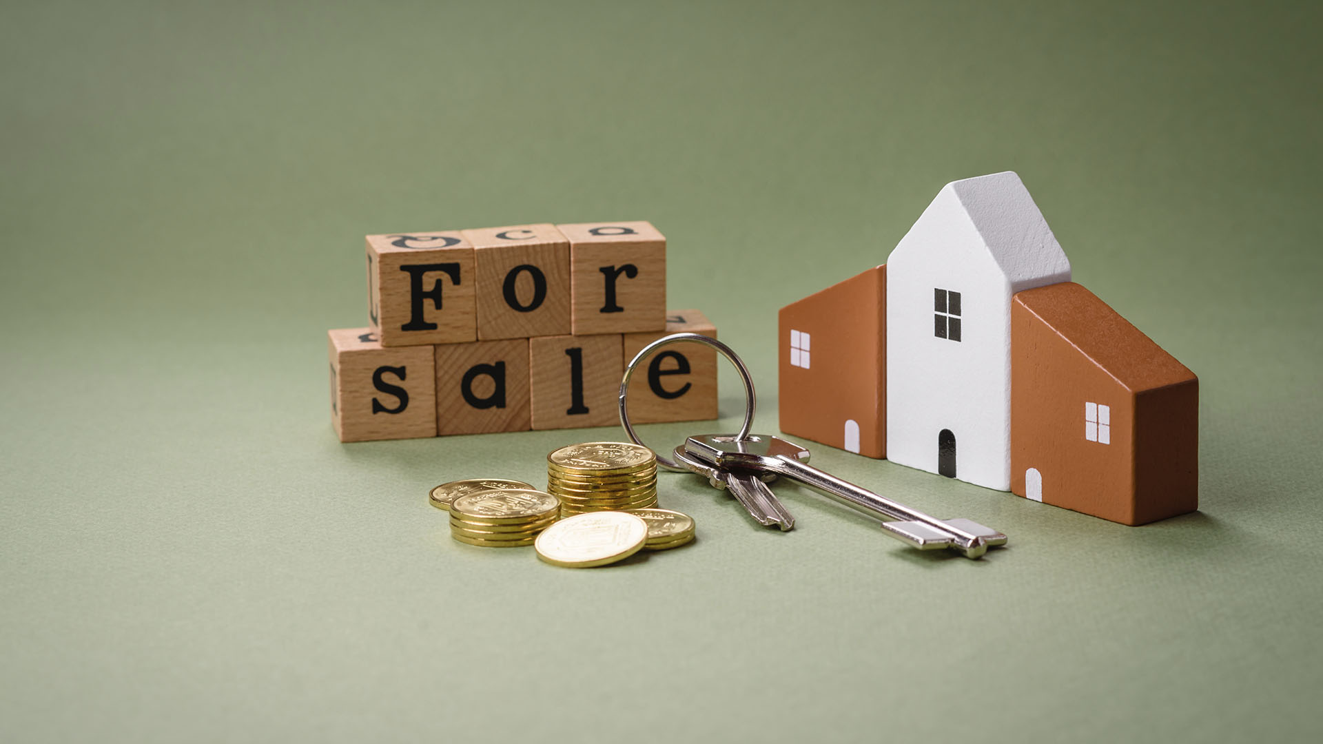 A model house, keys, stacks of coins, and wooden blocks spelling "FOR SALE" are arranged on a green background. Is renting ruining your future?