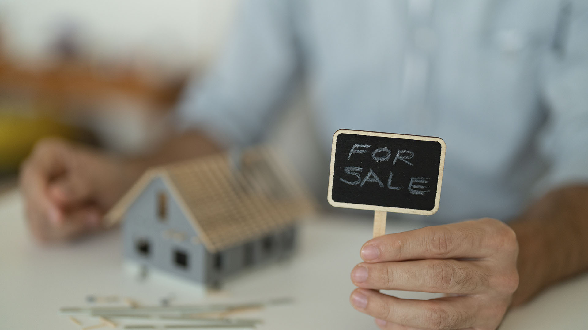 A person holds a small chalkboard sign that reads "For Sale" next to a miniature house model on a table. Is renting ruining your future? This could be the perfect opportunity for you.