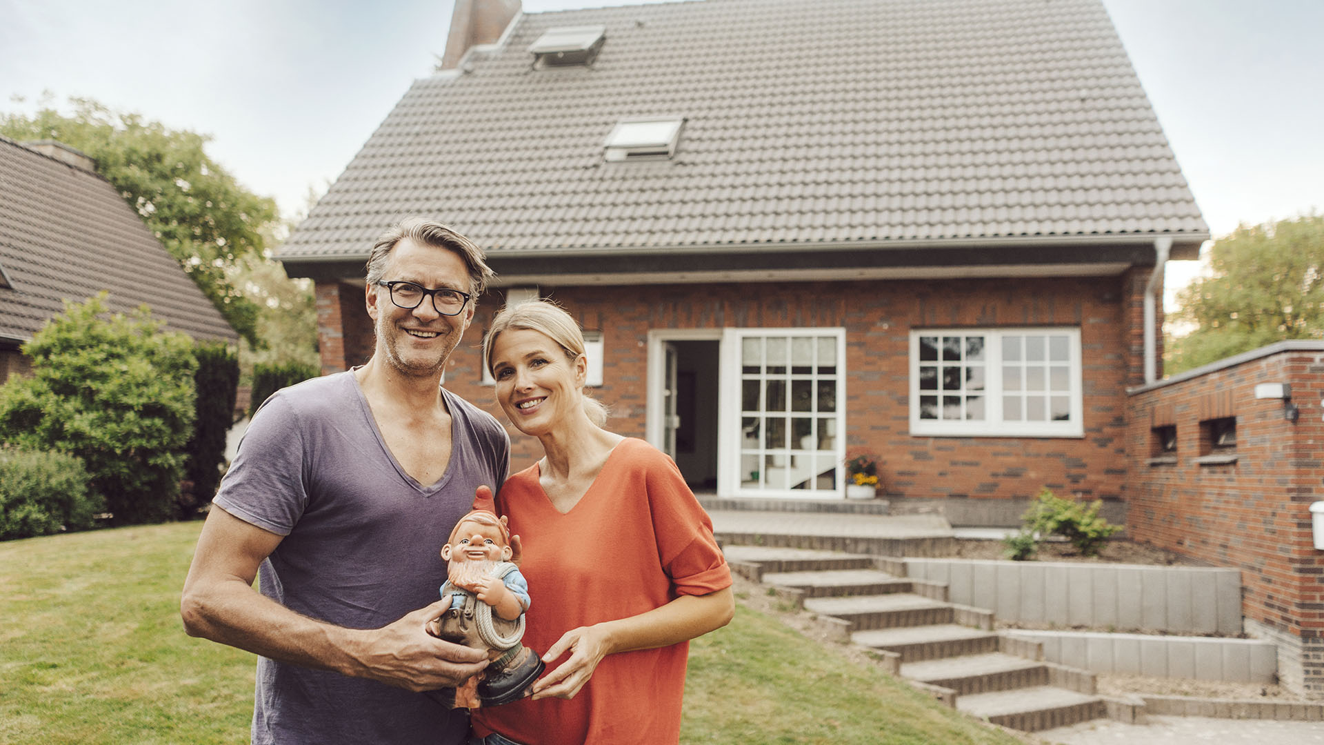A smiling couple stands in front of a brick house, holding a garden gnome. The man wears glasses and a purple shirt, while the woman is in a red top. They seem thrilled to be homeowners, no longer wondering if renting is ruining their future. The house has a well-kept lawn and shrubs.