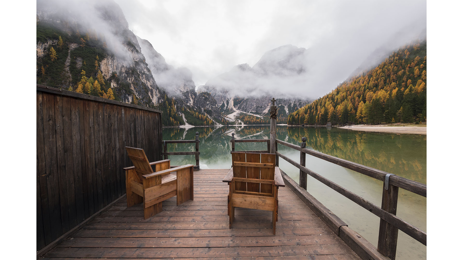 Two wooden chairs on a deck overlook a calm mountain lake, surrounded by mist-covered mountains and autumnal trees. Enjoy the serene view while reflecting on life's big questions: is renting ruining your future, or is this peaceful haven the escape you truly need?