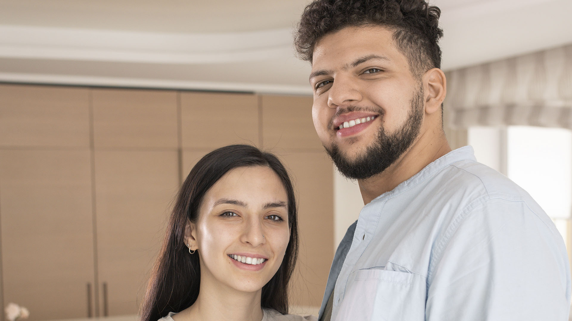A man and woman stand indoors, smiling at the camera. The background features light wood cabinets and a window with a curtain. Is renting ruining your future? Find out more as we explore their journey toward homeownership.