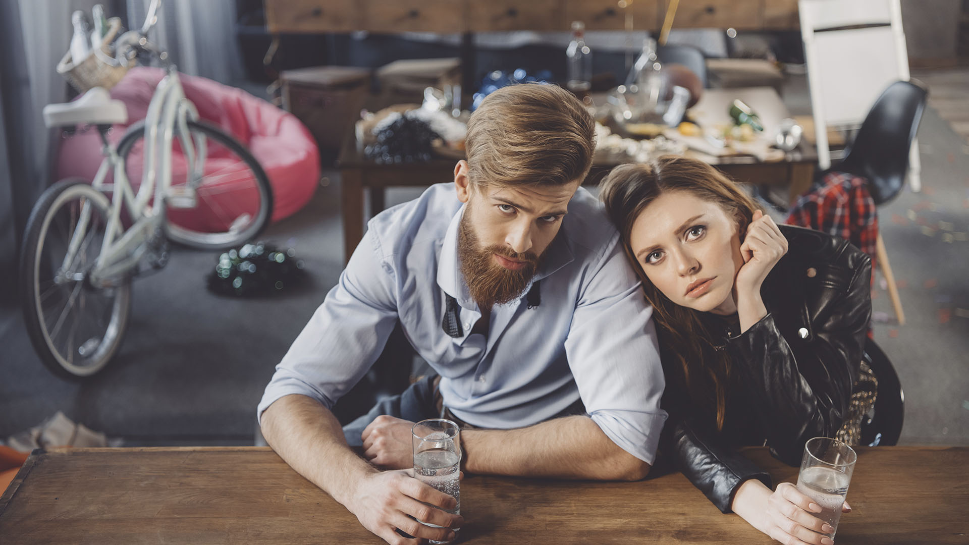 A man and woman sit at a table holding glasses of water, surrounded by a messy background that includes a bicycle, open bottles, and scattered objects. Is renting ruining your future?