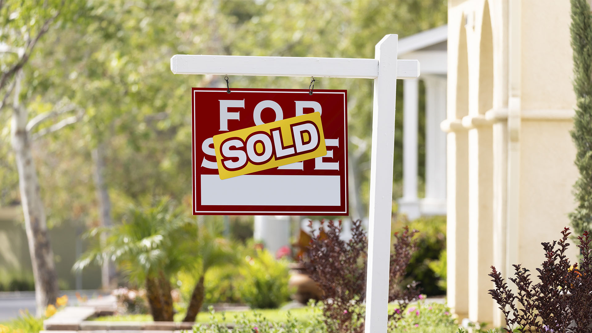 A "For Sale" sign with a "Sold" sticker is displayed in front of a house, indicating that it has been sold. Trees and foliage are in the background. Is renting ruining your future? This scene might just inspire you to take that next big step.