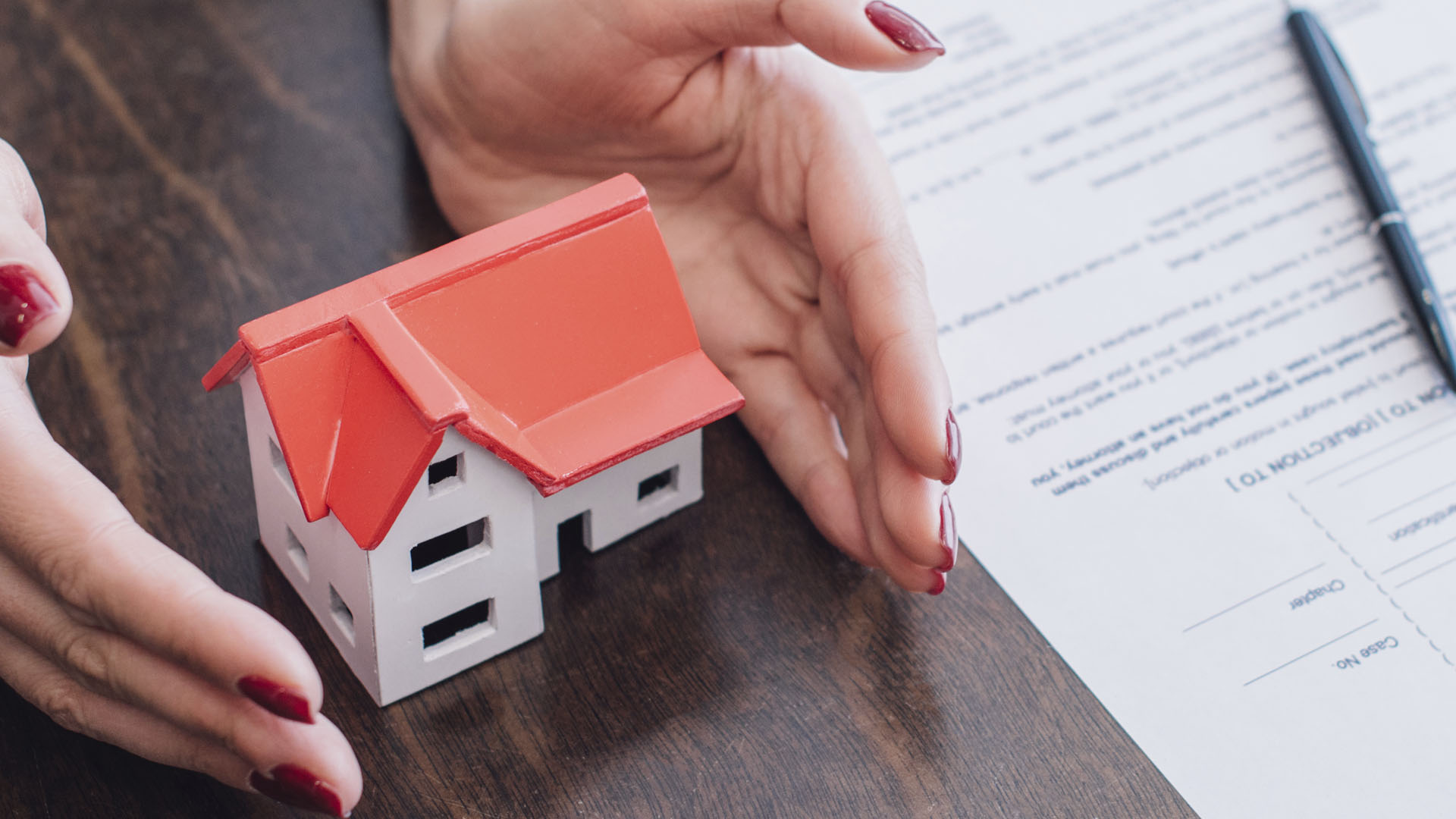 A person with red nails is holding a model house near a document and pen on a wooden table, prompting you to ask, "Is renting ruining your future?