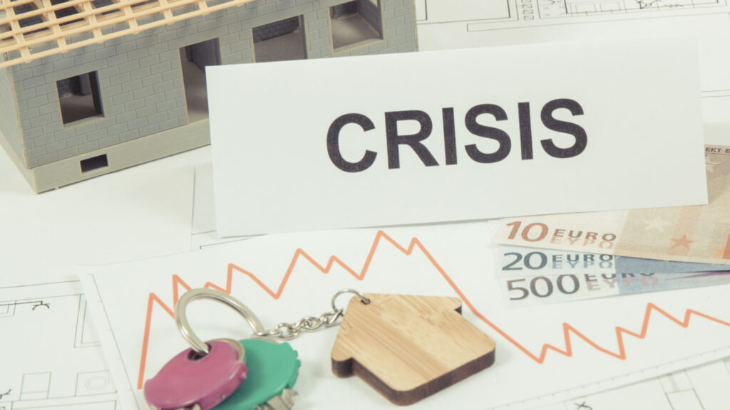 A house model, Euro banknotes, keys with a house-shaped keychain, and a sign reading "CRISIS" are placed on a table with a downward line graph—making you wonder, is renting ruining your future?
