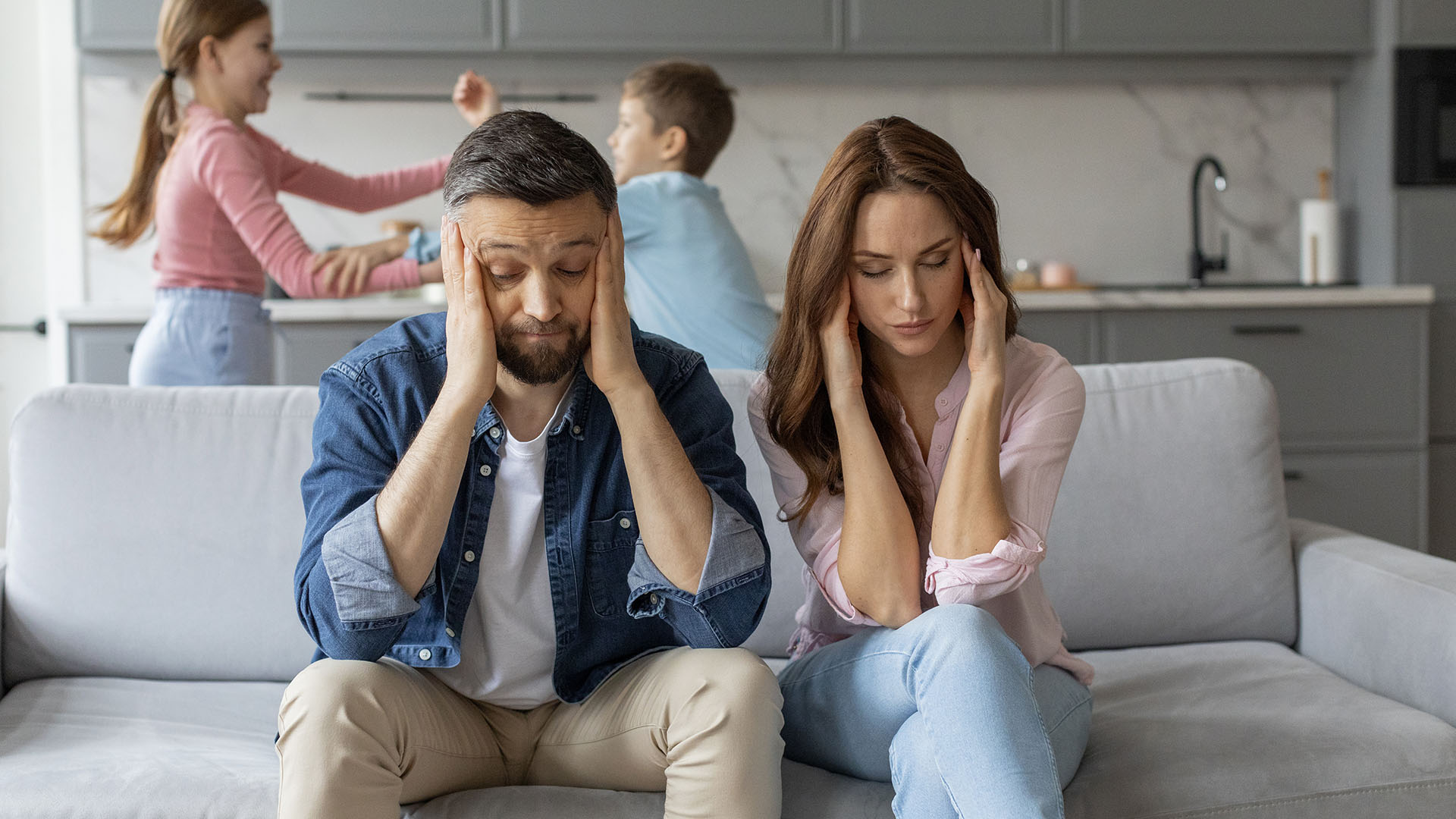A man and woman sit on a couch, covering their ears, while two children play energetically in the kitchen in the background. They ponder, "Is renting ruining our future?