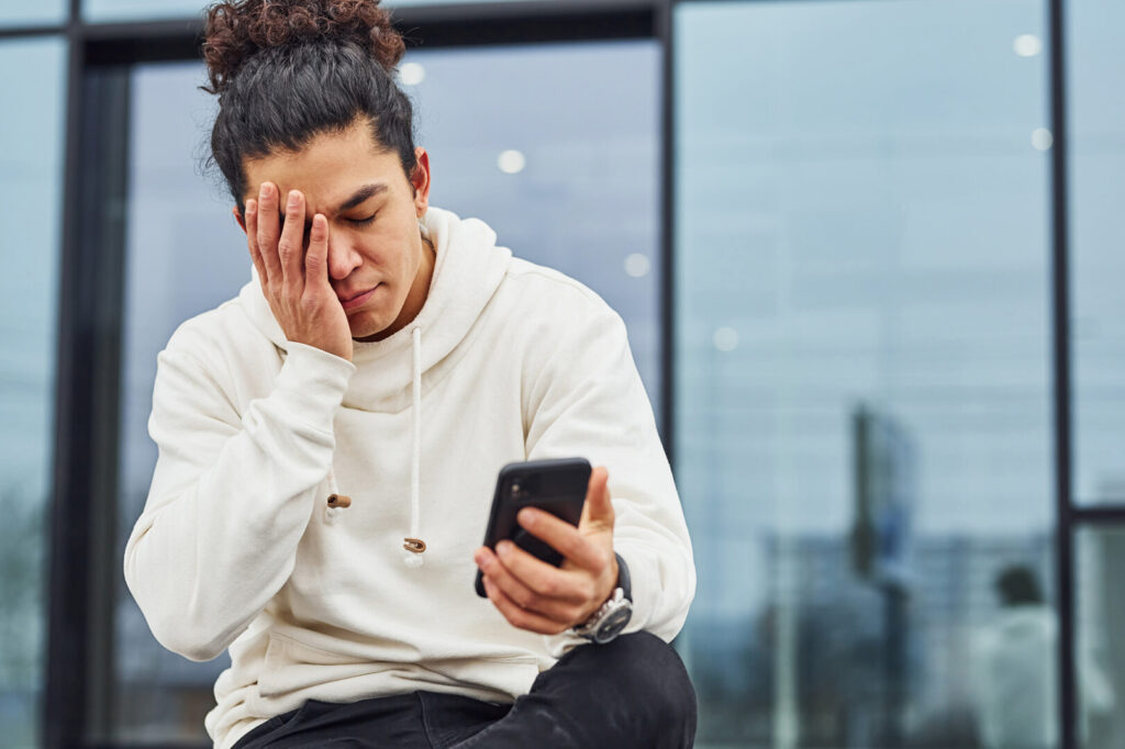 A person with curly hair, wearing a white hoodie, sits with one hand on their face while looking at a smartphone outside a modern glass building, seemingly lost in thought. Their expression reflects America's Mental Health Crisis as they grapple with reaching their breaking point.