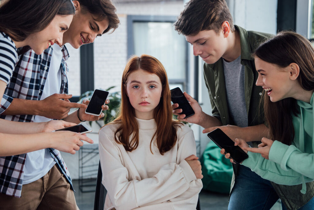 A girl sits with a distressed expression as five others stand around her, pointing and holding smartphones, capturing a moment emblematic of America's mental health crisis.