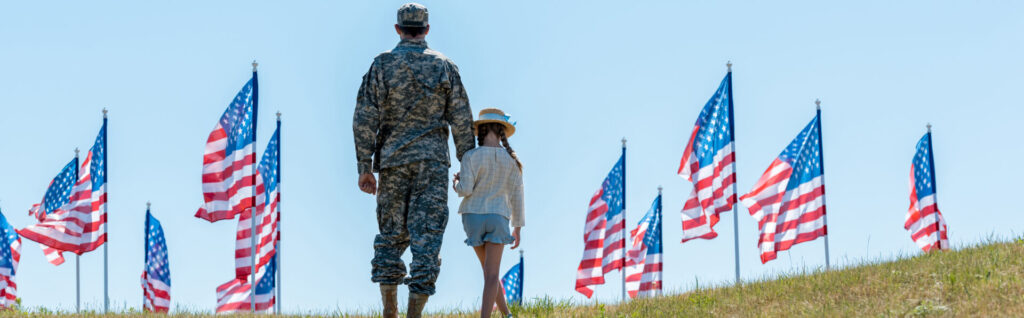 A person in military uniform stands holding hands with a child on a grassy hill, surrounded by numerous American flags fluttering against a clear blue sky, representing the dedication of Military Friendly® Companies supporting servicemembers and their families.
