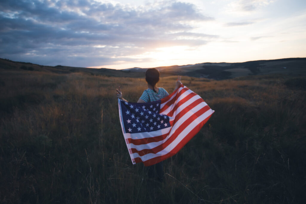 A person stands in a field at sunset, holding an American flag draped over their shoulders, with mountains visible in the distance—a tribute to our nation's top military companies.