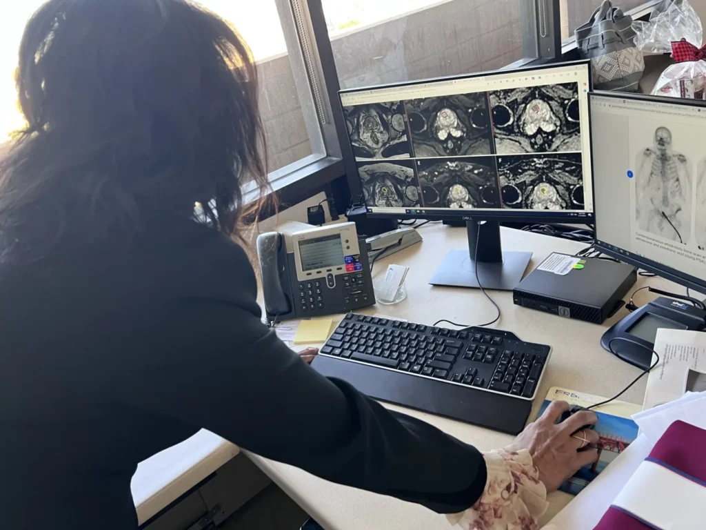 A person in a dark blazer examines medical imaging scans on a computer screen while standing at a cluttered desk, underscoring the significance of Men's Health matters amidst various items including a telephone and keyboard.