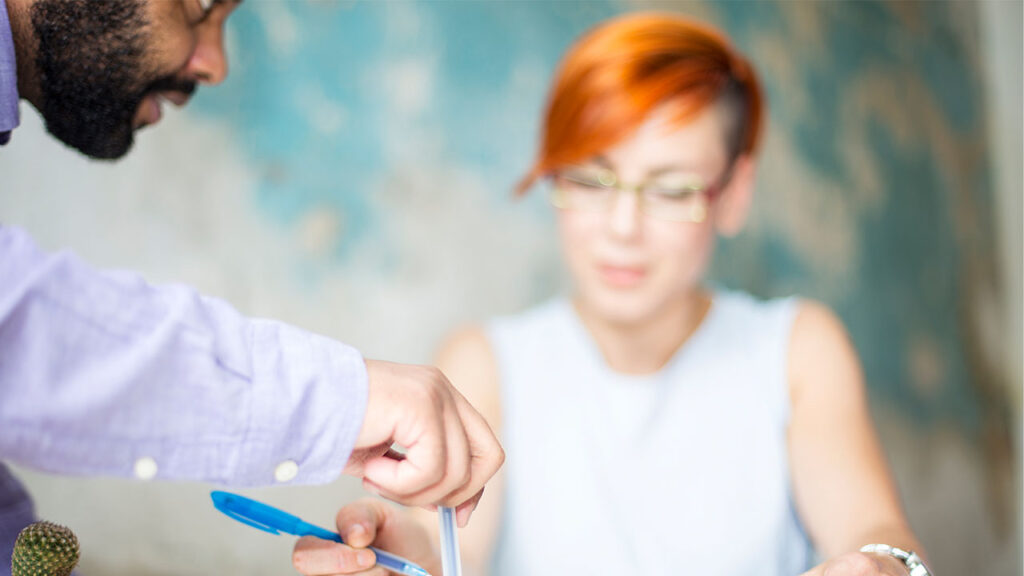         Two people collaborate at a table. One person, blurred in the background, observes while the other points to a document with a pen. Their discussion centers around changes in the job market due to the potential FTC noncompete ban.