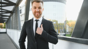 A man in a business suit stands on a bridge, giving a thumbs-up gesture. Behind him, the modern building with reflective windows mirrors the optimism of the improving job market on this bright September 4th.