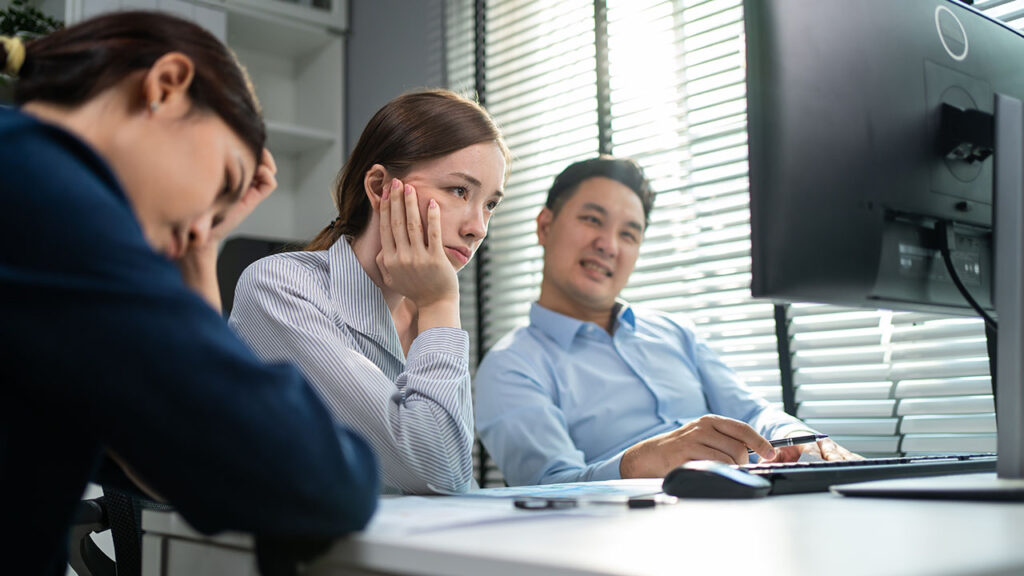 Two women and one man are sitting at a desk, looking at a computer screen; both women appear to be focused or thoughtful, while the man is smiling. The air is buzzing with excitement as they discuss the impending FTC Noncompete Ban set to revolutionize the job market.