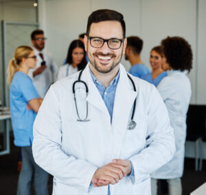 A smiling doctor with glasses and a stethoscope stands in front of a group of healthcare professionals having a discussion about men's health.