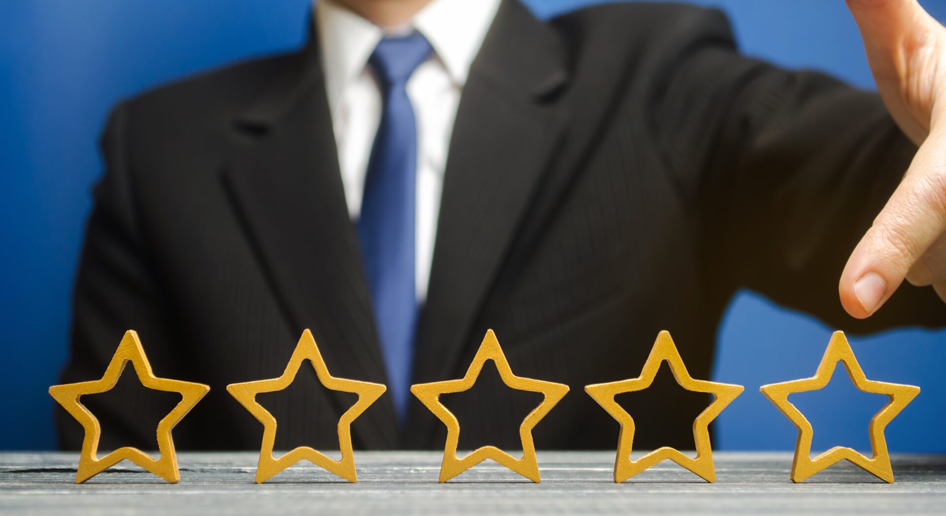 A person in a suit and tie is placing a fifth star in a row of four gold-colored star outlines on a wooden surface, against a blue background. Fake reviews got you down? This signifies genuine excellence.