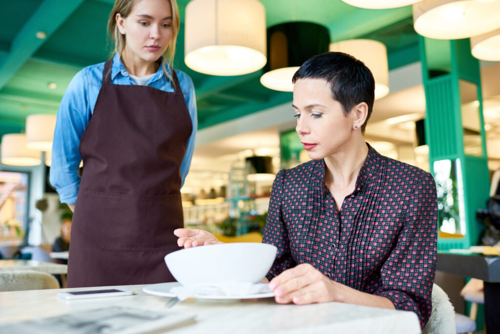 A woman in a patterned shirt looks at a bowl in front of her while a waitress wearing a brown apron stands by the table, seemingly unbothered by the fake reviews that got you down.