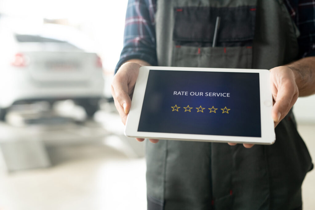 Person in work attire holds a tablet displaying a "Rate Our Service" screen with a five-star rating system. A vehicle can be seen in the blurred background. Fake Reviews Got You Down? Let genuine feedback brighten your day.