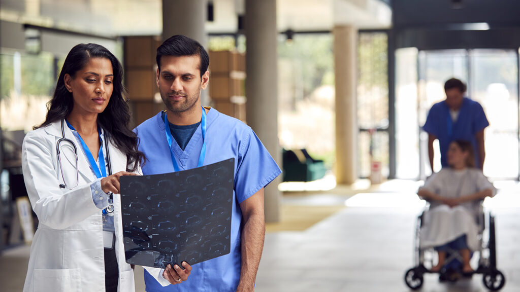 Two healthcare professionals examine an X-ray in a hospital hallway, emphasizing the importance of men's health. In the background, a person in a wheelchair and their caregiver offer a glimpse into the comprehensive care provided at the VA.