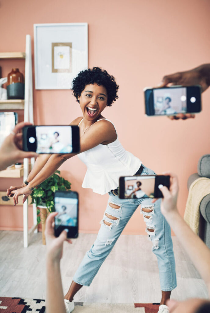 A person poses playfully while multiple individuals take photos of them with smartphones in a living room setting. Hold on to your TikTok moments—this playful snapshot is one for the memories!