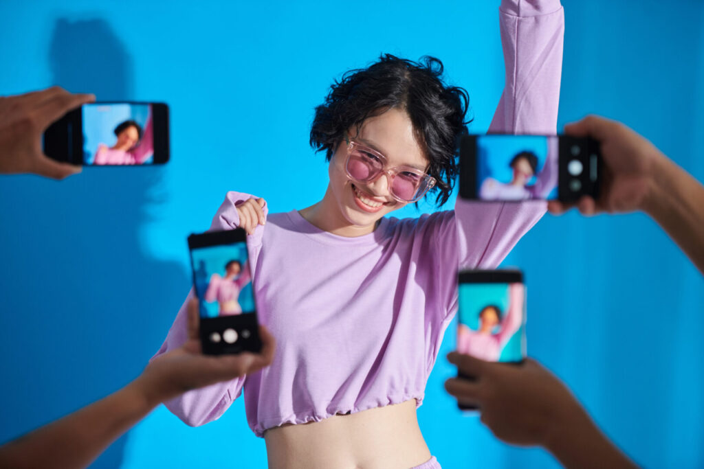 Person posing in a purple outfit and sunglasses in front of a blue background, while multiple hands hold smartphones taking pictures—hold on to your TikTok moments!